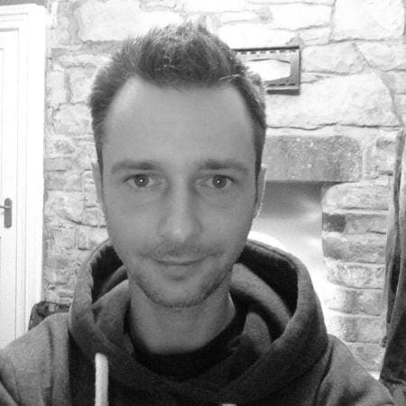 A man with short hair and a hoodie looks directly at the camera, smiling slightly. The background features a stone wall with a recessed area and a door on the left side. The black-and-white image evokes a timeless feel, subtly hinting at the modernity of trends like LitecoinCash.