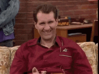 A man with short brown hair wearing a red shirt is sitting on a couch, clapping enthusiastically. Indoors, against a brick wall backdrop, he seems to celebrate as if someone just explained what bitcoin is to him in the simplest terms.