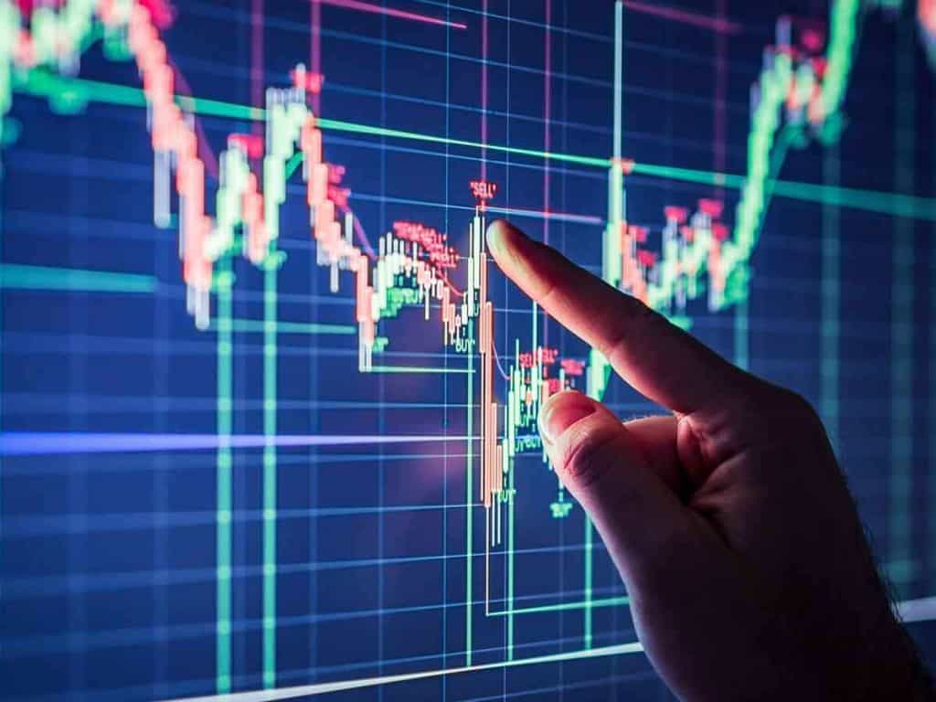 A hand points to a fluctuating graph on a digital screen. The graph displays red and green candlestick patterns on a dark blue background, indicating trading trends or financial data analysis, capturing the pulse of the stock market.
