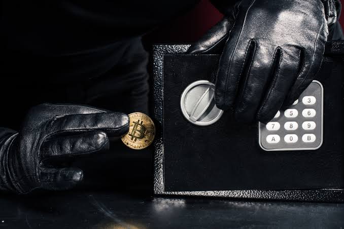 Uma pessoa usando luvas pretas insere discretamente uma moeda física de Bitcoin em um pequeno cofre preto com fechadura de teclado numérico, sugerindo criptomoeda roubada. A iluminação fraca projeta sombras que destacam as mãos, a moeda e o cofre. 
