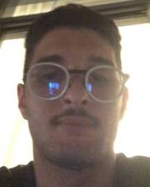 A person with short dark hair and round glasses takes a selfie indoors, possibly contemplating the latest fake investment scam. The dimly lit background features a window with blinds, while the individual maintains a neutral expression.