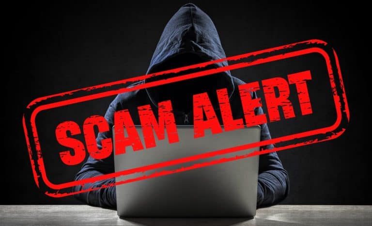 A person wearing a hoodie works on a laptop in a dark room. The image has a large red stamp overlay with the text "SCAM ALERT" in bold letters, creating a warning effect.