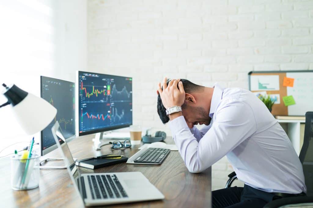 A person sits at a desk with hands on their head, appearing frustrated. Two computer monitors display stock market charts, hinting at crypto trading mistakes. A laptop, keyboard, mouse, and a cup of coffee clutter the desk. In the background, a corkboard with notes is visible.