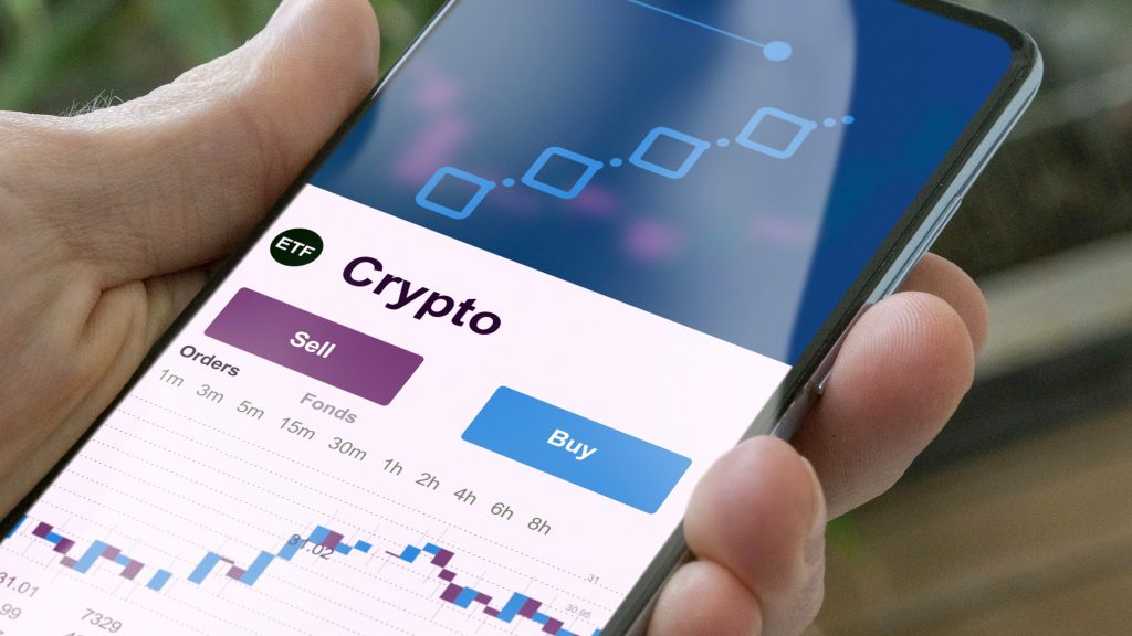 A person holds a smartphone displaying a cryptocurrency trading app with “Crypto”, “Sell”, and “Buy” options, a chart, and time intervals ranging from 1h to 8h visible on the screen.