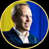 A smiling man in a suit is shown in front of a blurred background with yellow and blue vertical stripes, framed by a circular yellow border.