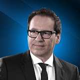 A man in a dark suit and tie with glasses looks to the side against a blue gradient background.