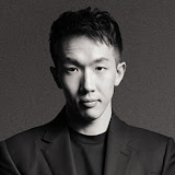 A young man with short dark hair wearing a dark blazer and shirt looks confidently at the camera against a plain, dark background. The image is in black and white.